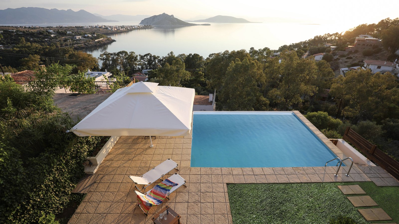 View of Eucalyptus Villa's terrace with outdoors dinner table, sun beds and the infinity swimming pool. In the back you can enjoy a panoramic view of the western part of the Saronic gulf and the islands of Moni and Agistri.